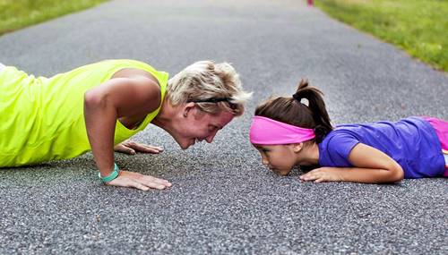 older  woman  and  child  facing  each  other  smiling  as  they  do  push  ups  on  tarmac  floor